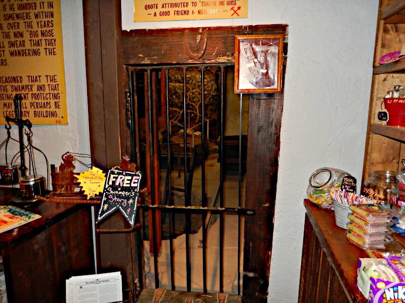 Interior view of a rustic store or saloon featuring a wooden doorway with iron bars resembling a jail cell. Surrounding it are various signs, decorative items, and shelves stocked with colorful merchandise, creating an eclectic old-west themed atmosphere.